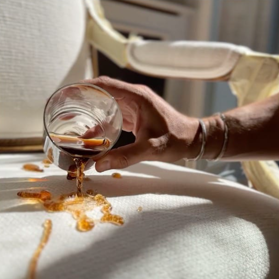 Mano derramando bebida oscura de un vaso sobre una superficie texturizada blanca.