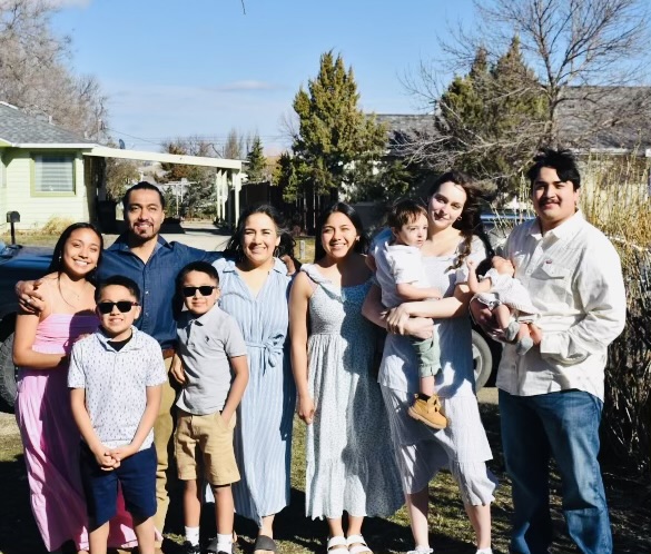 Una familia de nueve miembros, todos sonrientes, de pie al aire libre en un día soleado; tanto los niños como los adultos llevan ropa veraniega informal.