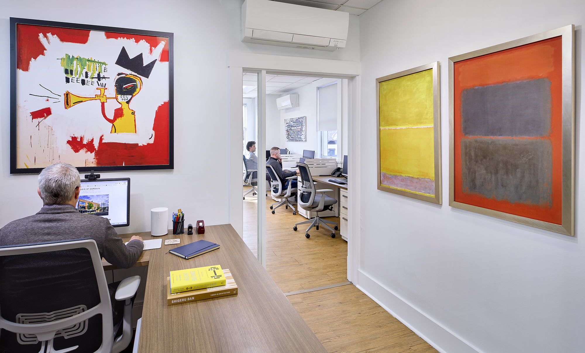Modern office space showing a man with gray hair seated at a desk in the foreground working on a computer, with paintings on the walls including a colorful abstract artwork with a trumpet and crown, and two framed abstract color block paintings. Through a doorway, two other people sit at desks with computers. The room has wooden floors, white walls, and air conditioning units mounted above.