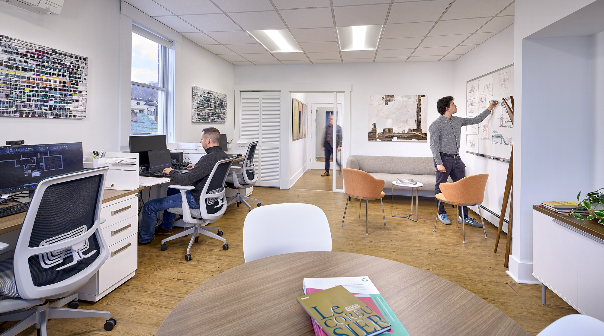 Modern office space with wooden floor, a man sitting at a desk with multiple computer monitors on the left, another man writing on a whiteboard on the right, and a gray couch with two orange chairs in the background; round table with books in the foreground.