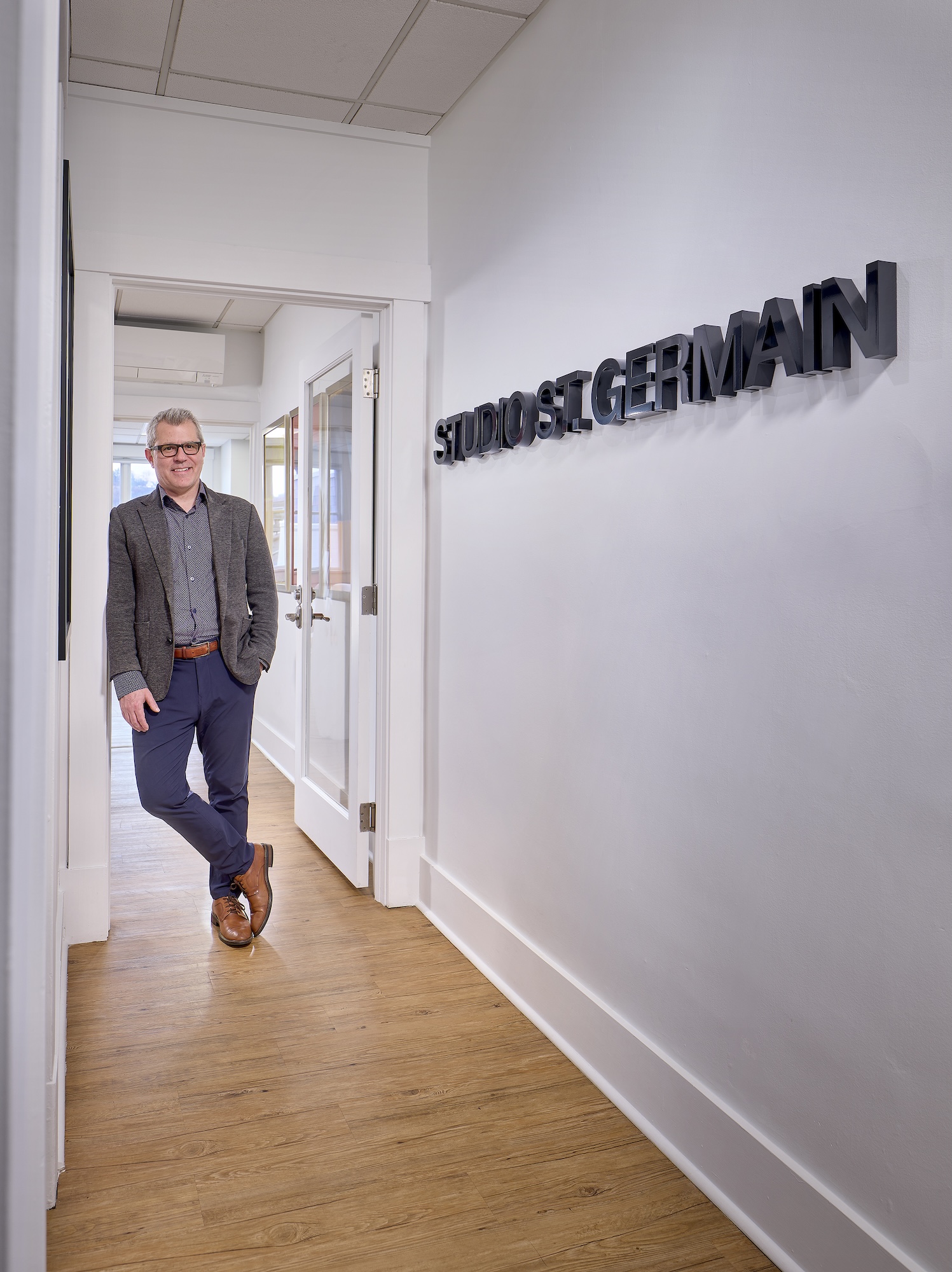 A man wearing glasses, a grey blazer, patterned shirt, blue pants, and brown shoes stands with his hands in his pockets, leaning against a door frame in a bright hallway with light wood flooring and white walls, next to a wall sign that reads 'STUDIO ST. GERMAIN'.