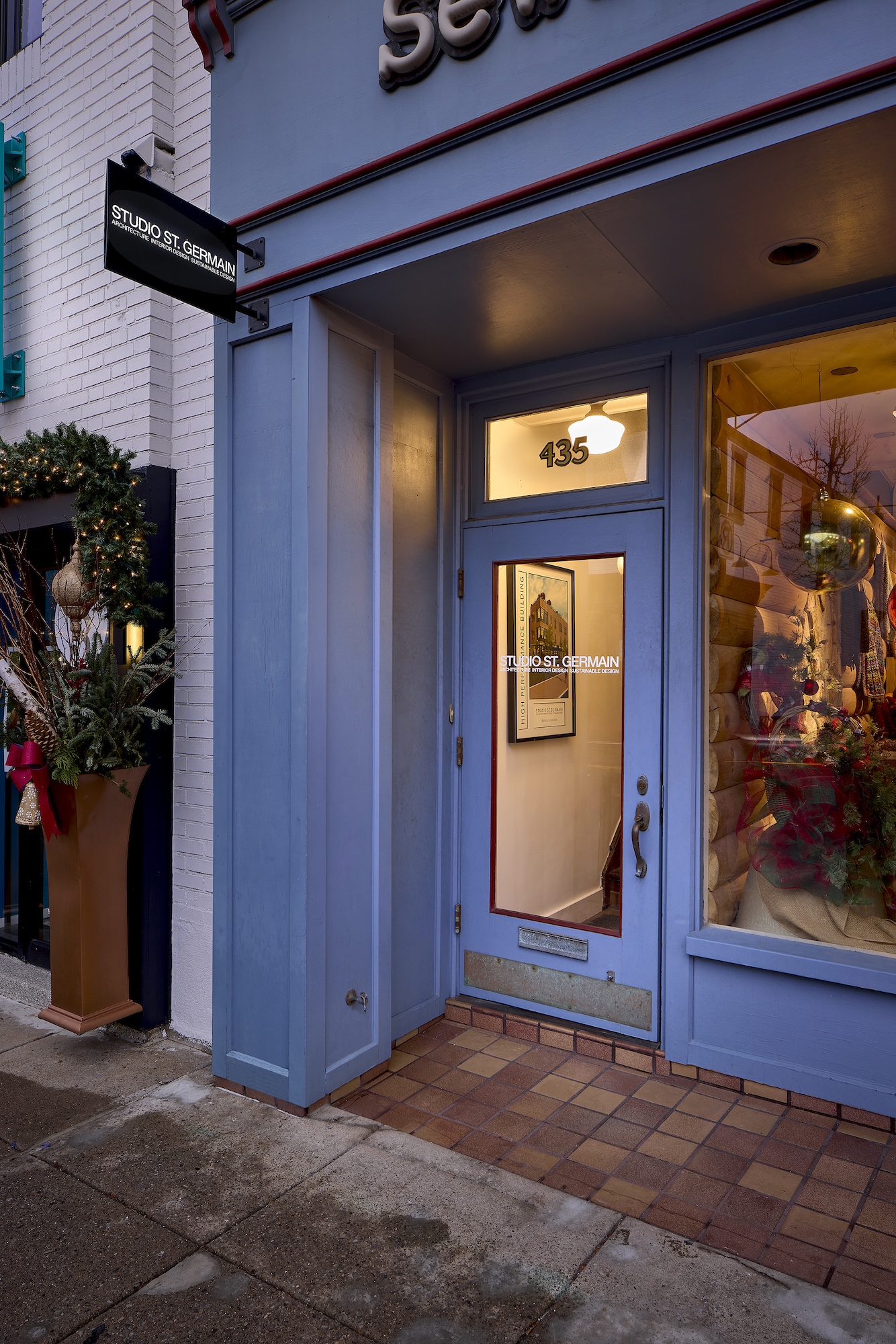 Entrance of Studio St. Germain with a blue door and frame, address number 435 above the door, and a glass panel showing a framed picture and interior lighting; next to the door is a window with holiday decorations including festive greenery, red ribbons, and ornaments; a black sign with white text for Studio St. Germain hangs from the building; a tall planter with greenery and red decorations stands on the sidewalk beside the entrance.