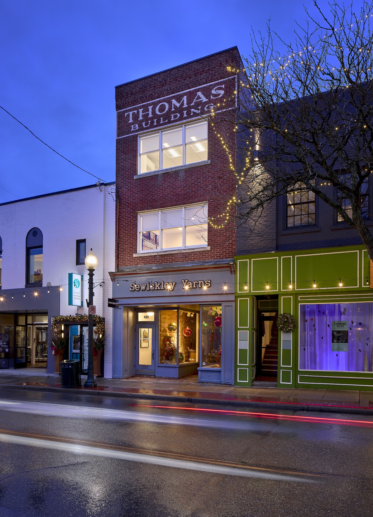 Street view at dusk of the brick Thomas Building with illuminated Sewickley Yarns shop on the ground floor and decorated tree with string lights nearby.