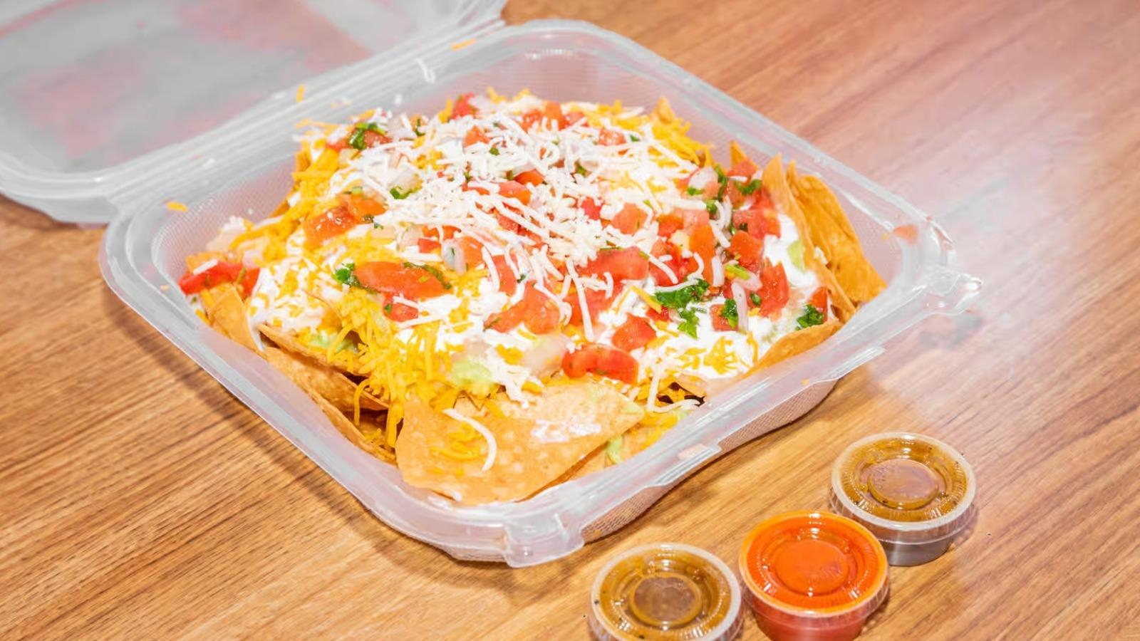 Takeout container of nachos topped with shredded cheese, diced tomatoes, cilantro, and sour cream, with three small containers of sauces on the side.