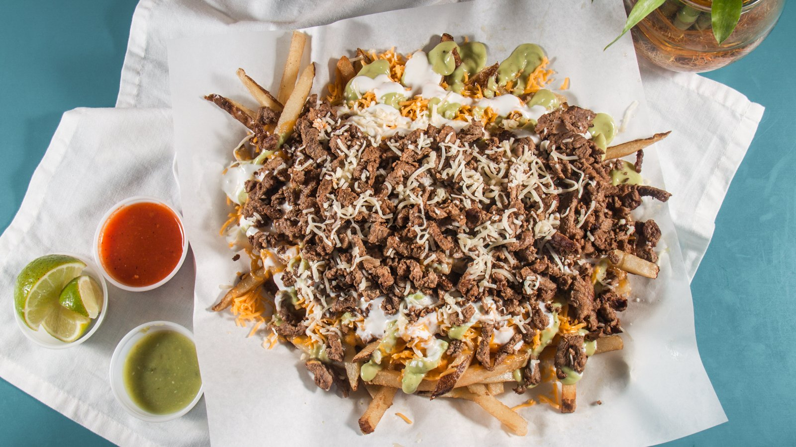 Loaded fries topped with seasoned beef, shredded cheese, and drizzled with green and white sauces, served with lime wedges and salsa on the side.