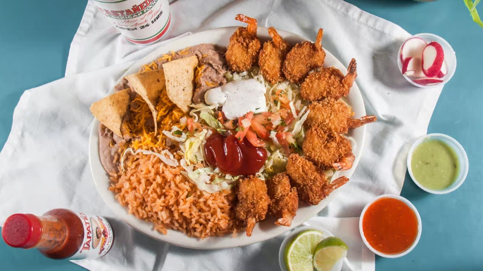 Plate with fried shrimp, Mexican rice, refried beans with chips, shredded lettuce and tomato topped with salsa and sour cream, surrounded by lime wedges, radish slices, and three sauces on a white cloth.