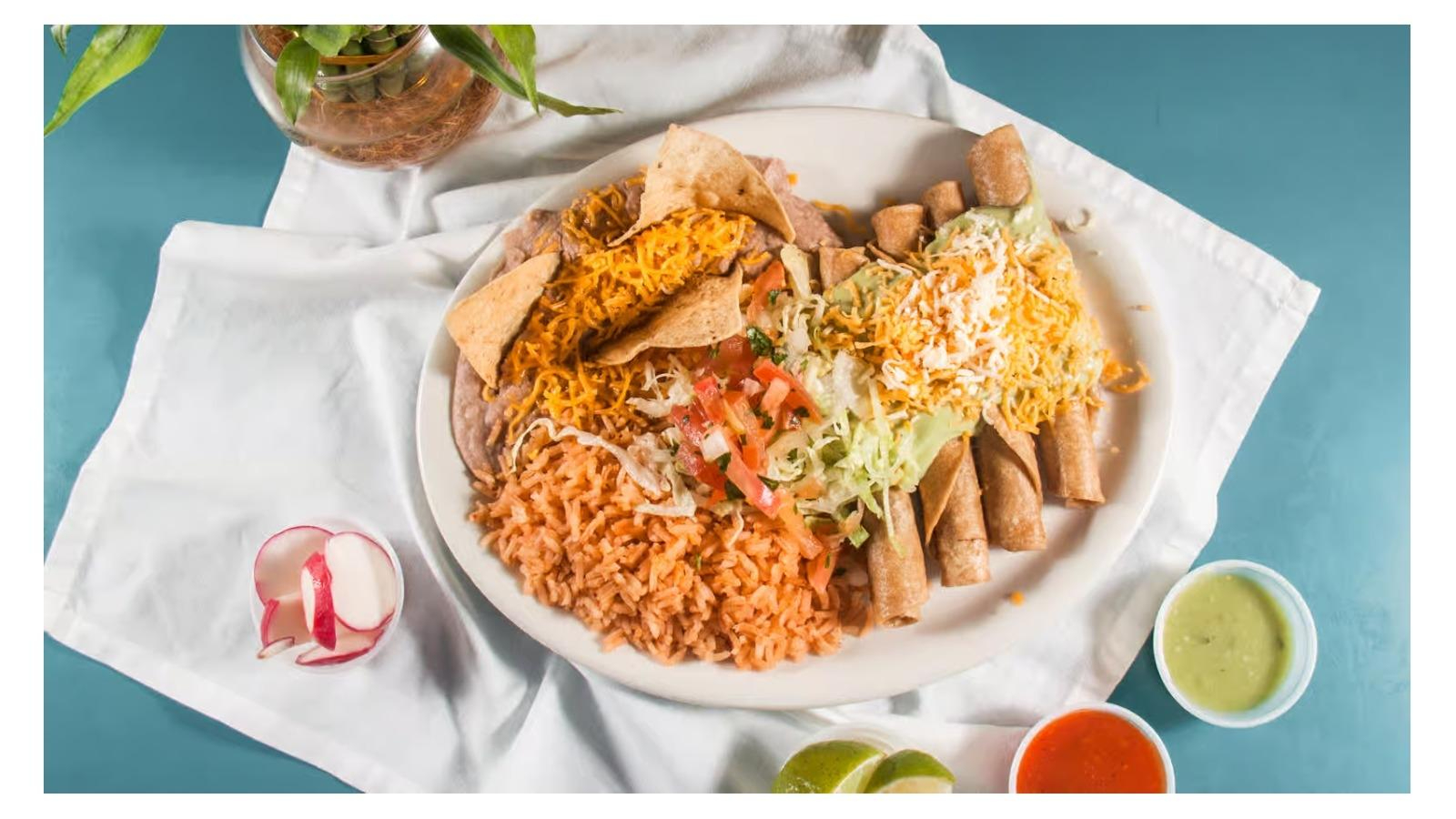 Plate with Mexican food including rice, refried beans topped with cheese and chips, rolled taquitos with lettuce, tomato, and shredded cheese, accompanied by sliced radishes, lime wedges, and cups of red and green sauces on a white cloth and blue surface.