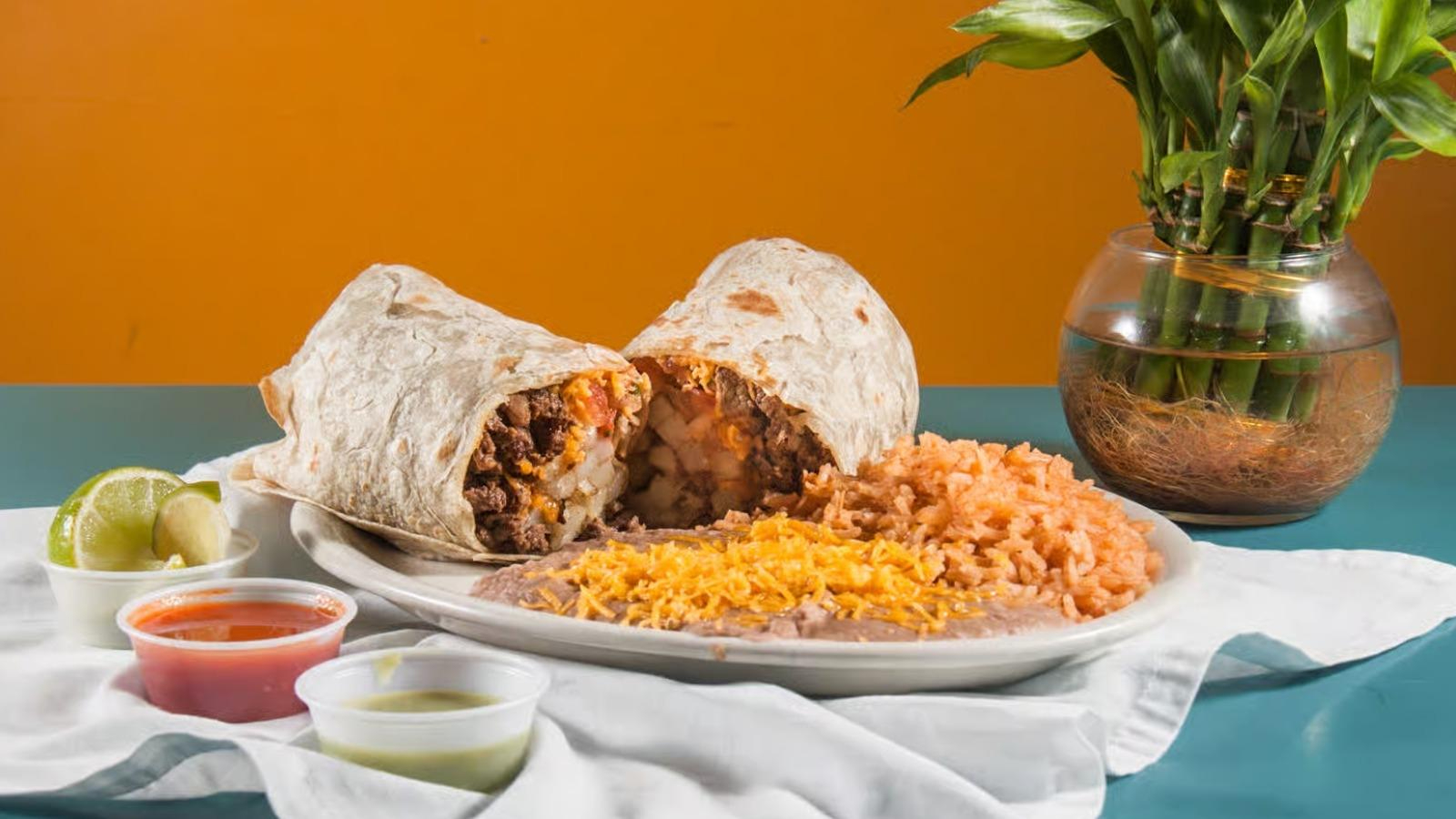 Plate with a halved beef burrito, Mexican rice, refried beans topped with shredded cheese, lime wedges, and two small containers of salsa and green sauce.