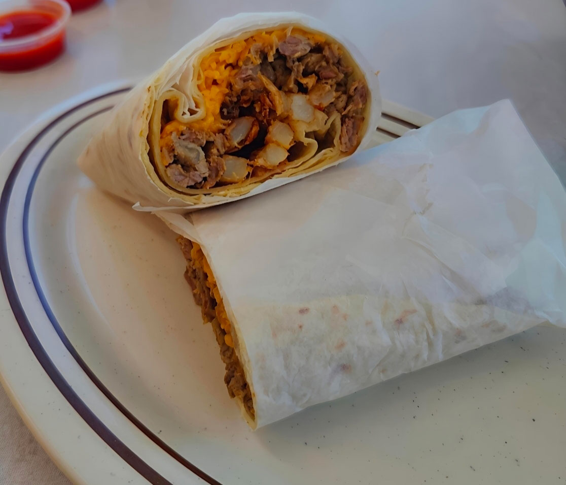 Half-wrapped burrito cut in half on a white plate with meat, diced potatoes, and shredded cheese inside.