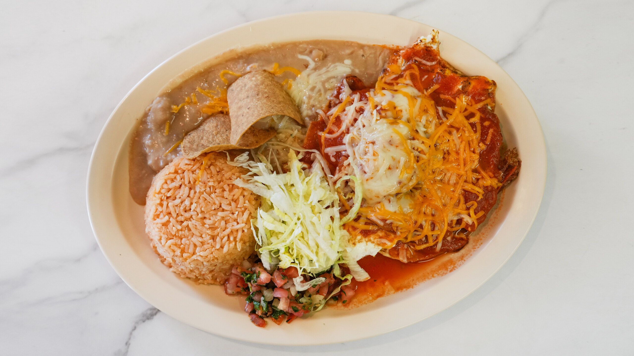 Mexican plate with rice, refried beans topped with tortilla chips and cheese, shredded lettuce, pico de gallo, and an enchilada covered in melted cheese and red sauce.