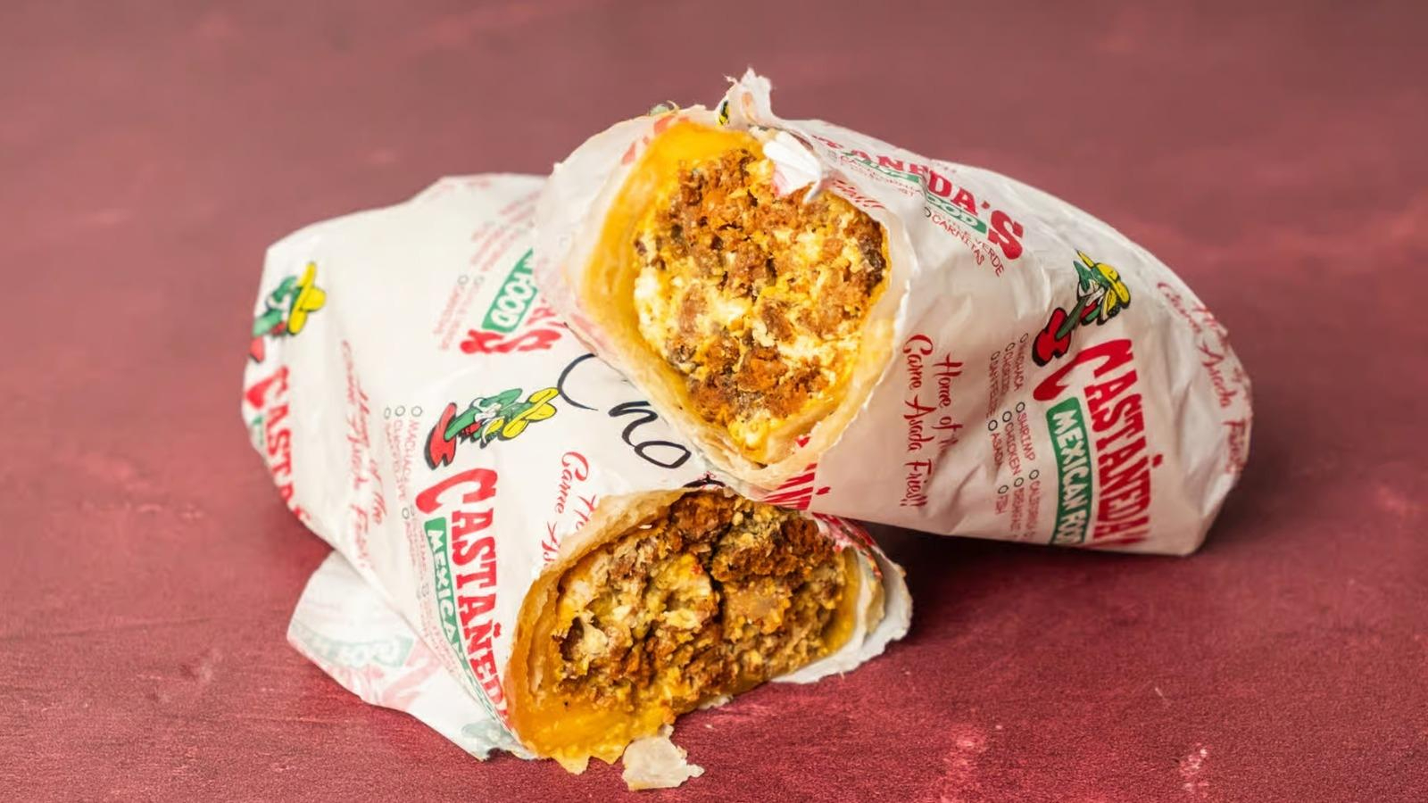 Half of a breakfast burrito filled with scrambled eggs and chorizo sausage wrapped in branded white paper on a reddish surface.