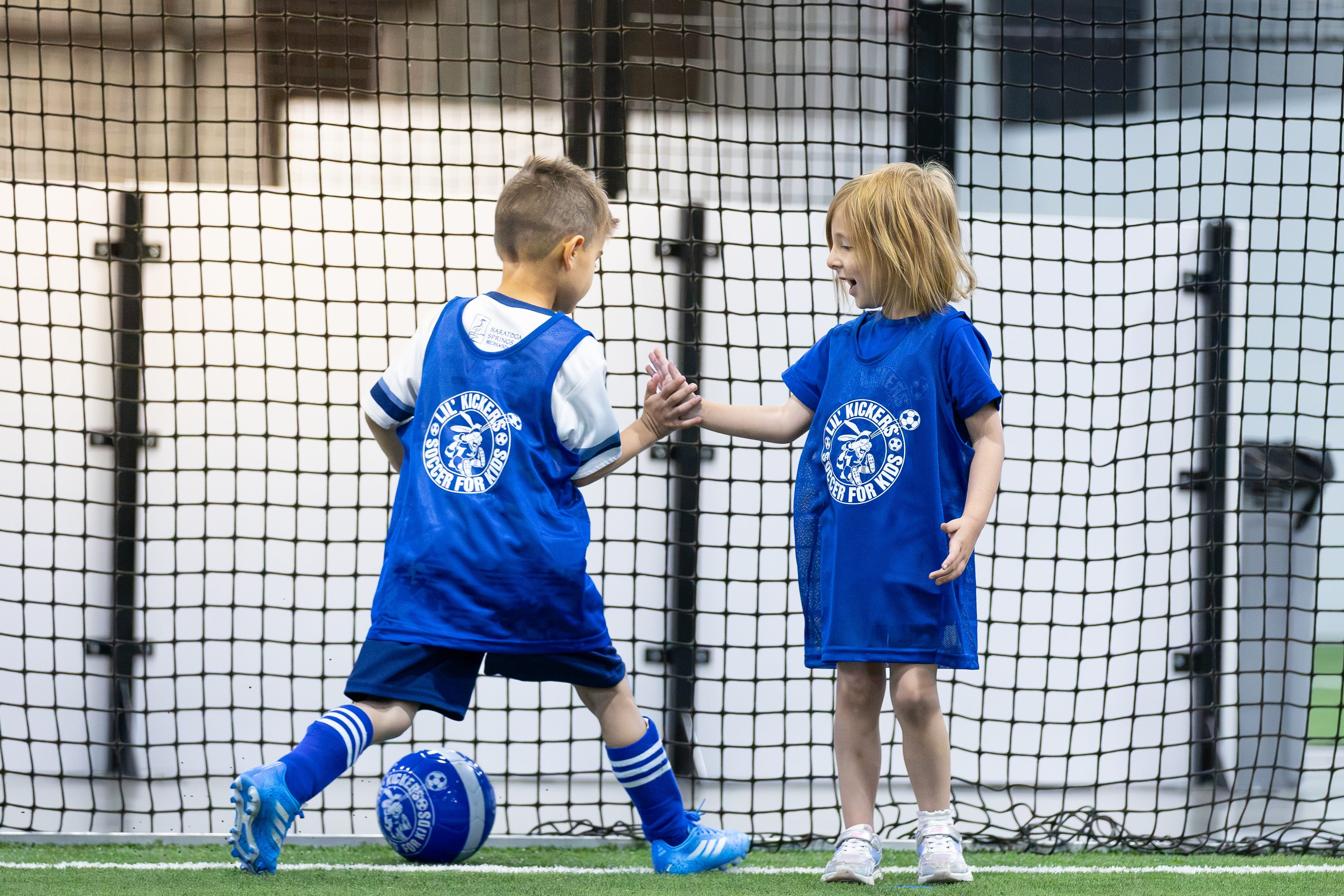 Two Lil' Kickers players in blue pinnies on an indoor turf field