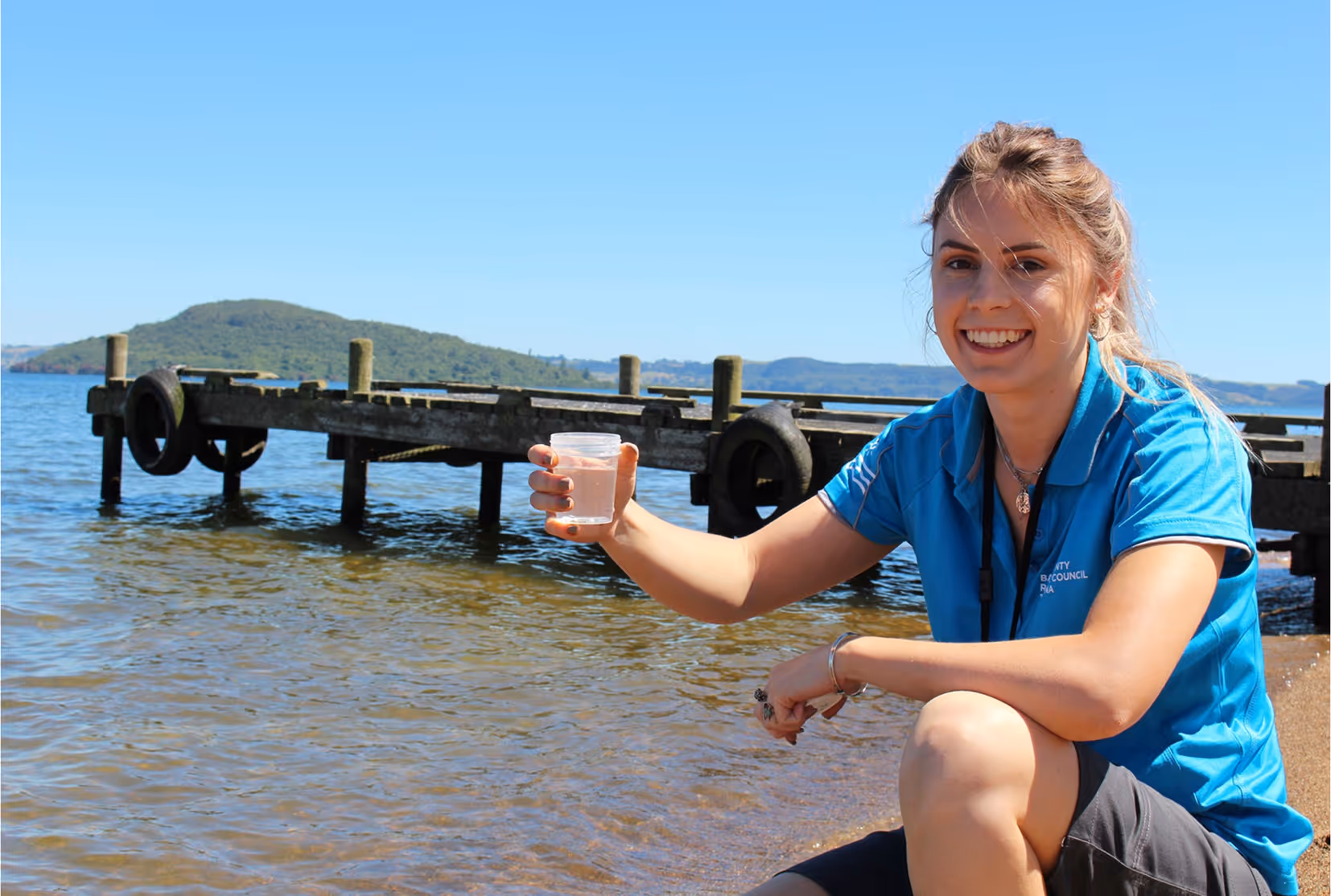 Bay of Plenty Regional Council staff member crouching by the lakeside holding a water sample container, with a wooden jetty and hills in the background