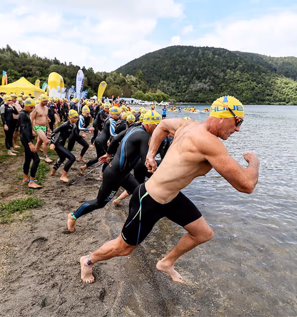Open-water swimmers in yellow caps racing into a Rotorua lake at the start of a swimming event, with bush-clad hills in the background