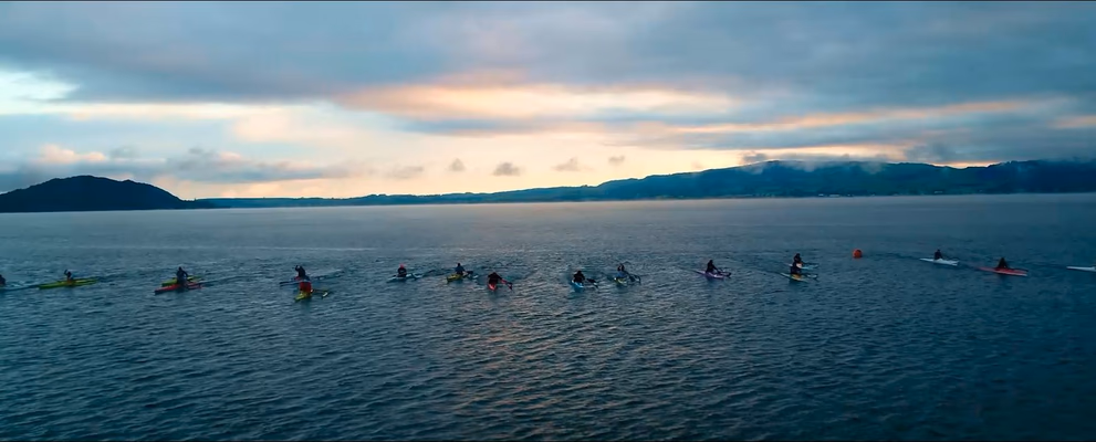 Aerial view of waka ama paddlers on a Rotorua lake at sunrise, with Mokoia Island and surrounding hills in the background