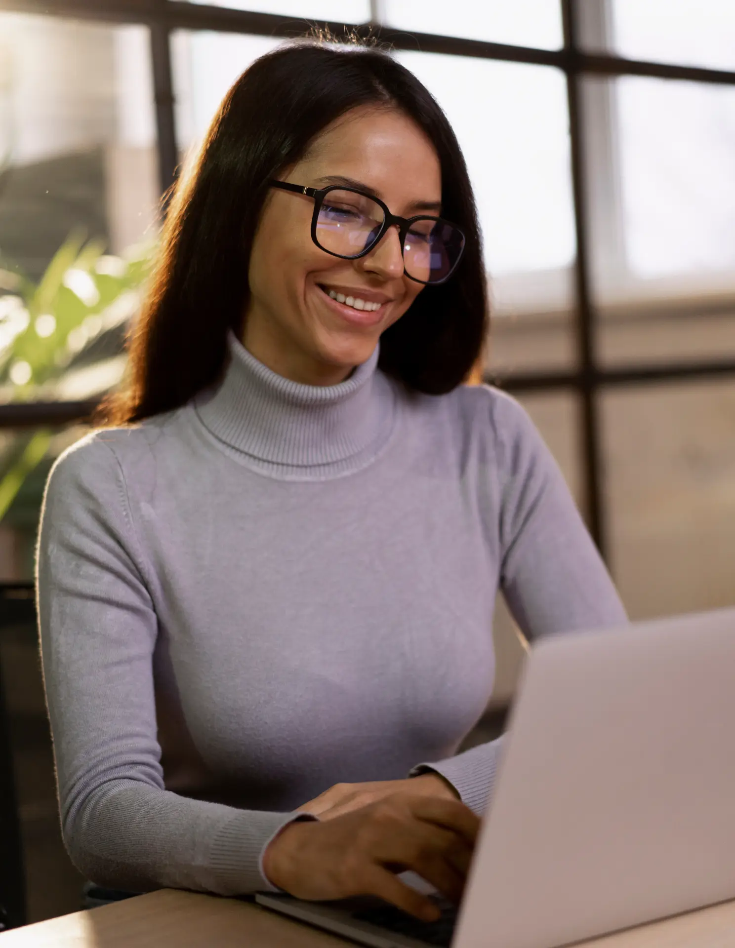 Smiling woman with glasses typing on a laptop in a bright room.