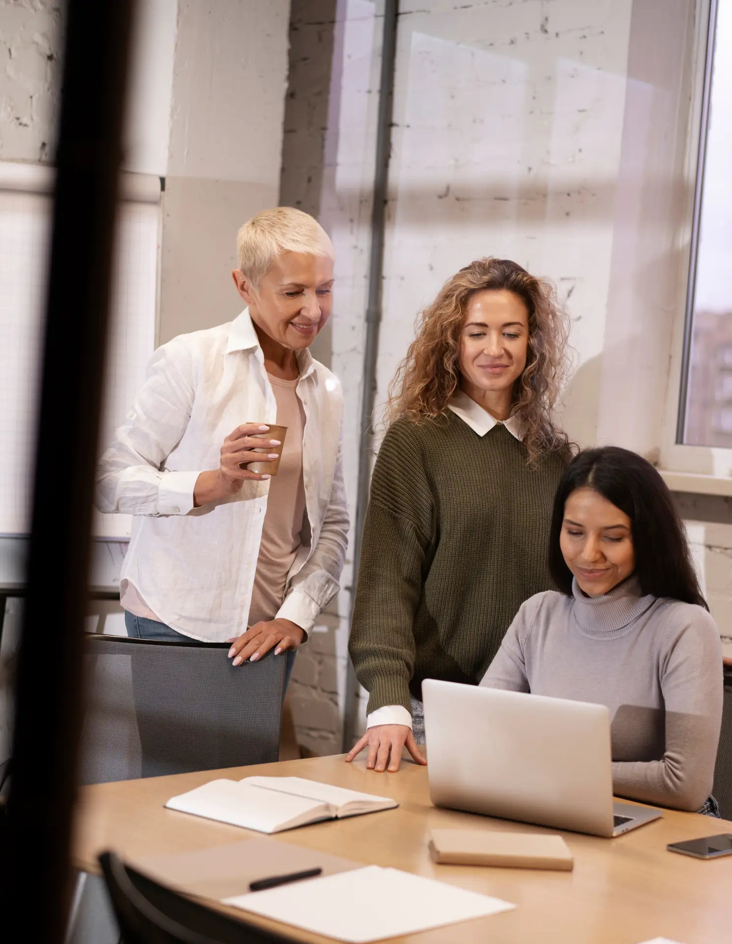 Three women in a meeting room, one sitting and working on a laptop while the other two stand nearby, one holding a coffee cup.