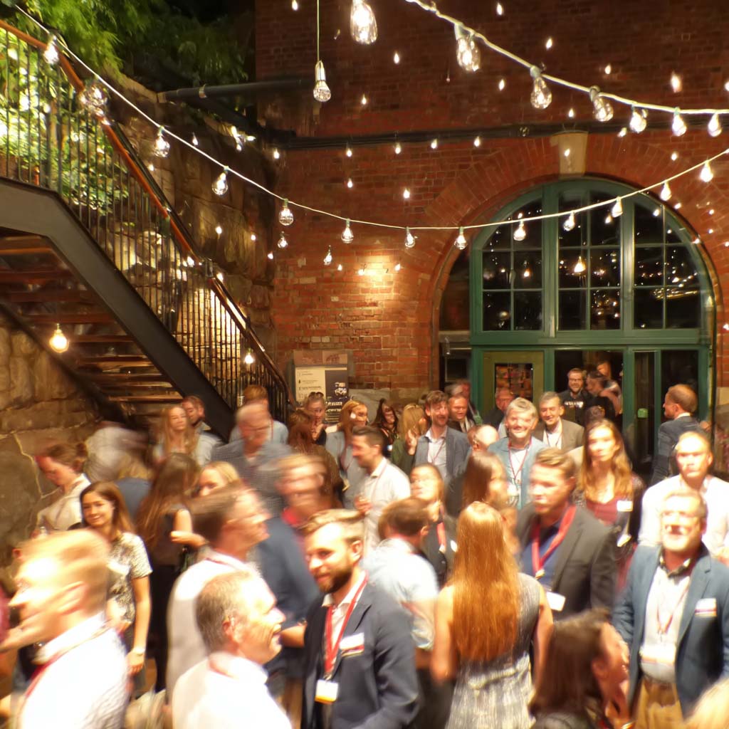 Crowd of people socializing at an indoor event with string lights and brick walls.