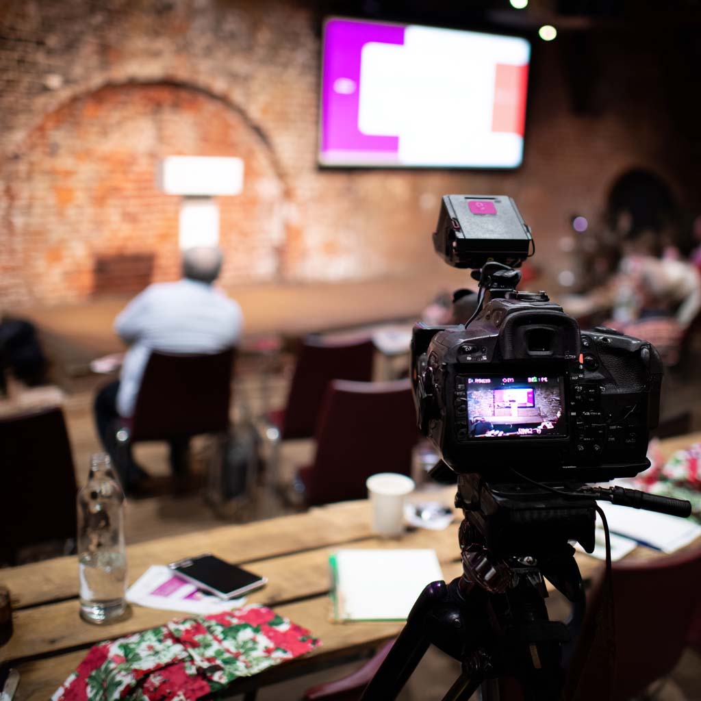 Camera on a tripod recording a person sitting in front of a brick wall with a blurred screen in a live event setting.