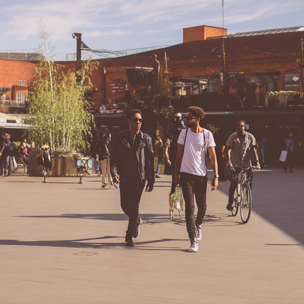 Two men walking on a sunny urban plaza with one man in a leather jacket and the other in a white t-shirt carrying groceries, while another man rides a bicycle behind them.