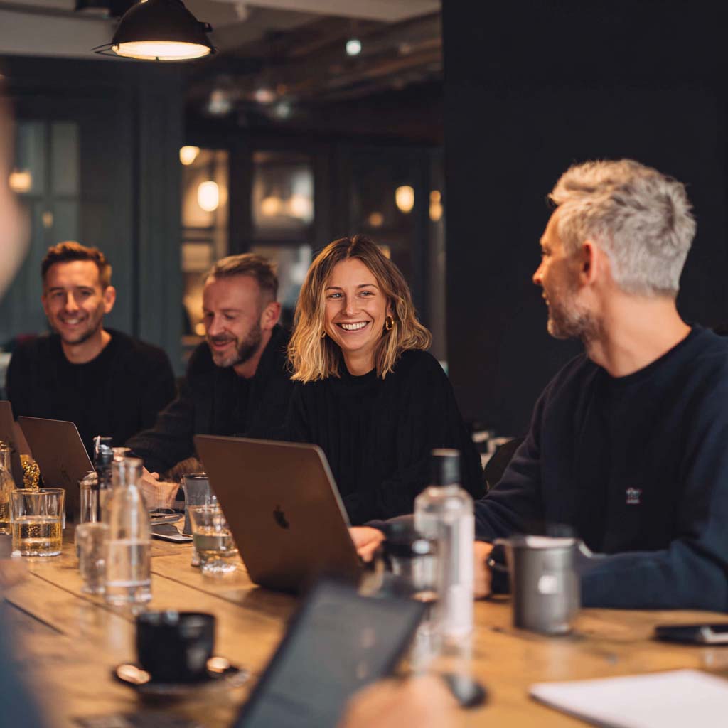 Four colleagues smiling and chatting around a wooden table with laptops and drinks in a cozy office setting.