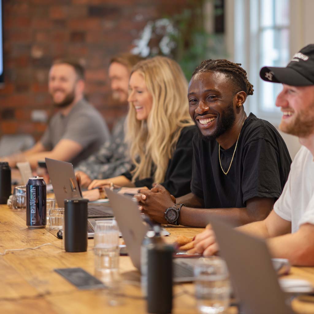 A diverse group of smiling young professionals working together at a wooden table with laptops and drinks.