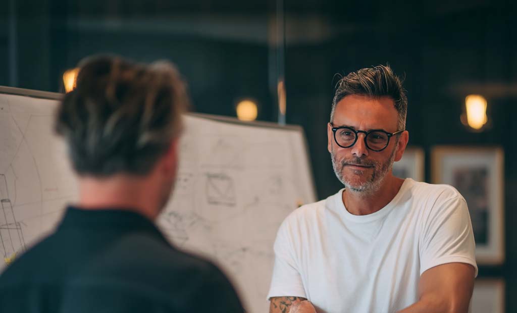 Two men engaged in discussion near a whiteboard with sketches and diagrams in an office setting.