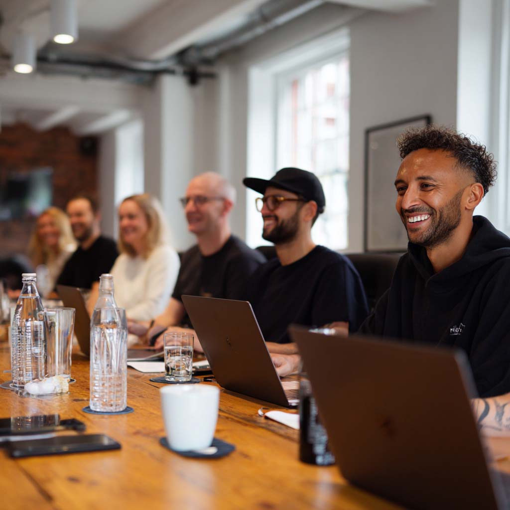 Group of six people smiling and sitting in a meeting room with laptops on a wooden table.