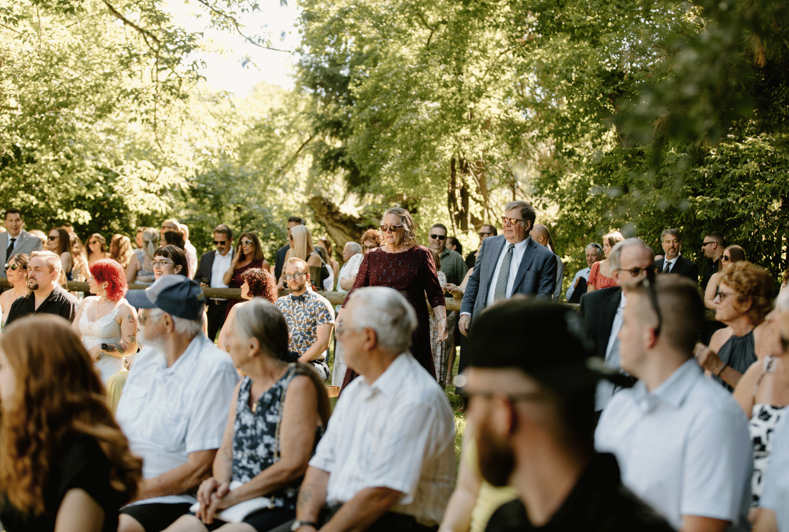 Outdoor gathering of diverse people seated and standing under trees on a sunny day.