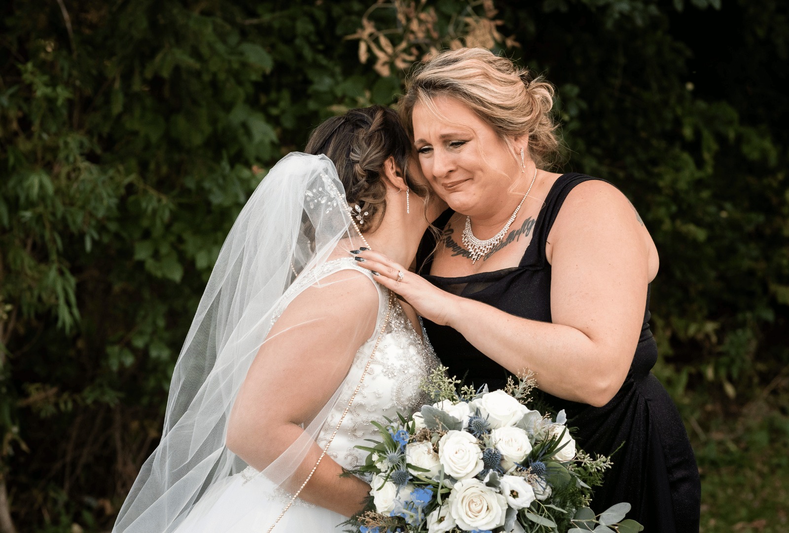 Bride in white veil and dress embraces a teary woman in black dress holding a bouquet of white flowers.