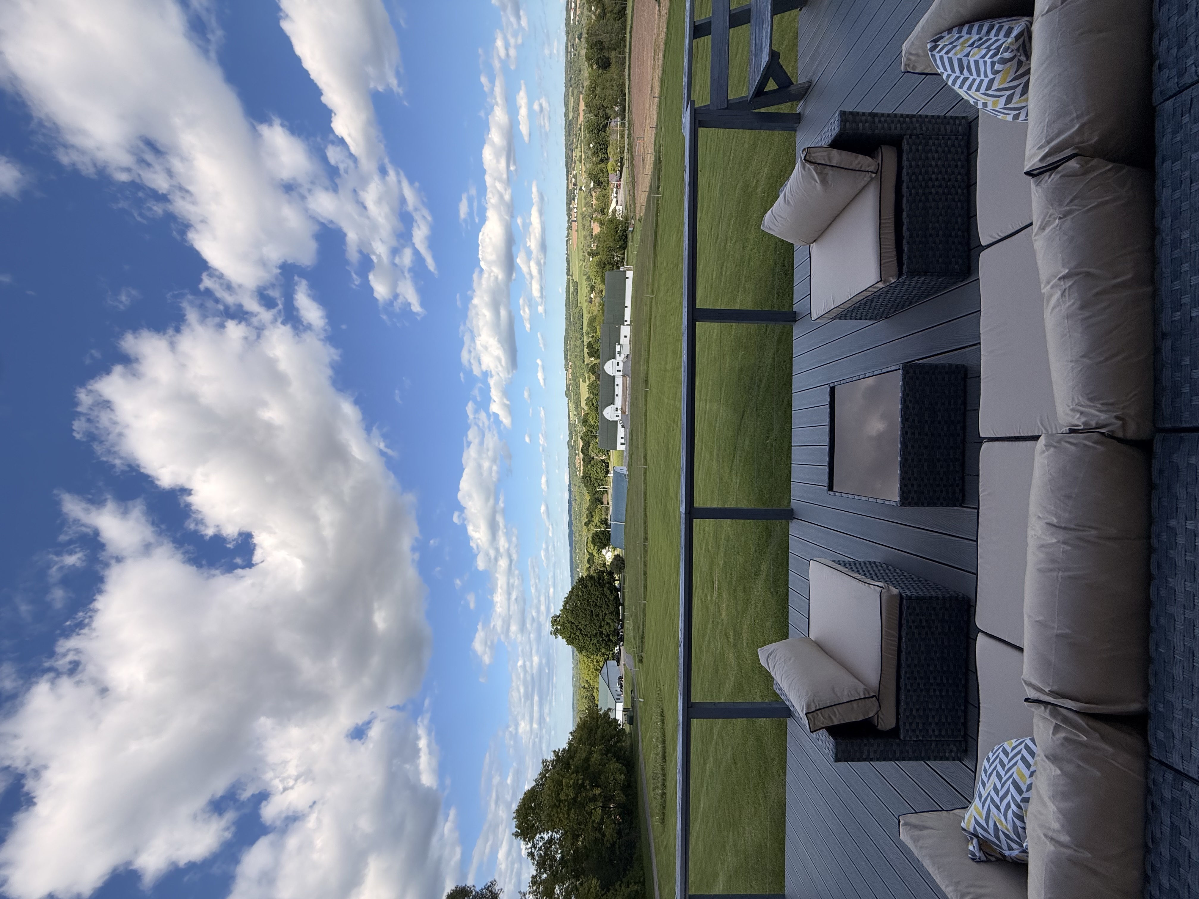 Outdoor deck seating with cushions overlooking green fields, white barns, and a blue sky with clouds.