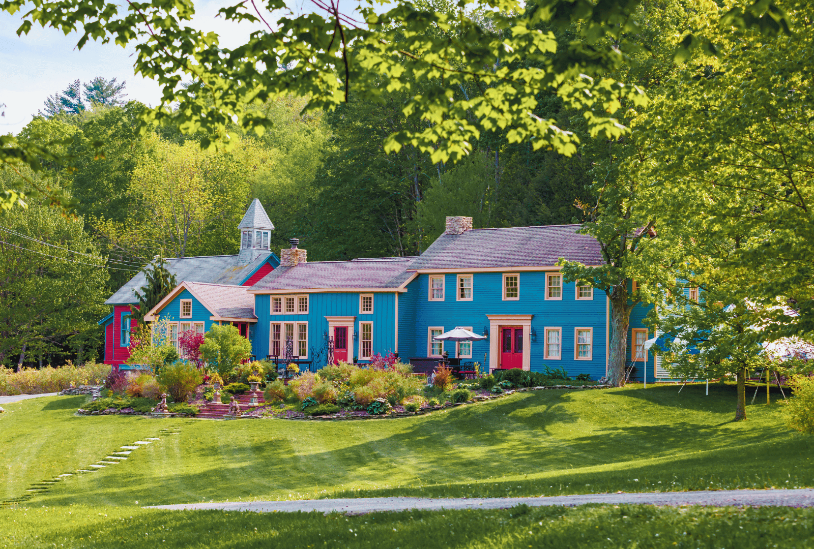 Blue house with red doors set in a lush green yard surrounded by trees and colorful shrubs.