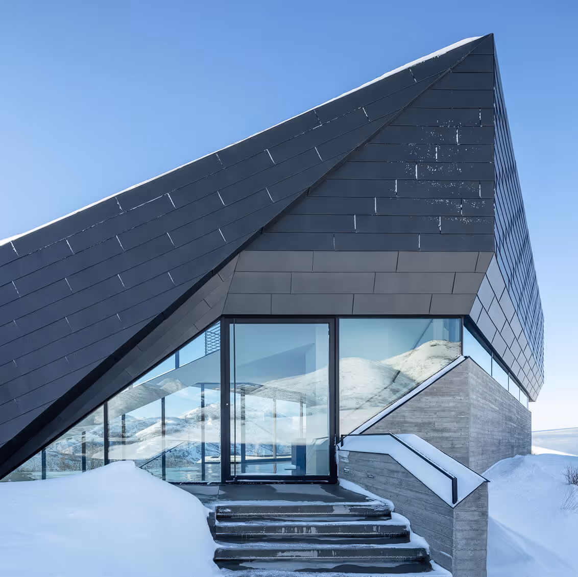 Modern angular building with large glass windows and snow-covered stairs in a snowy landscape.