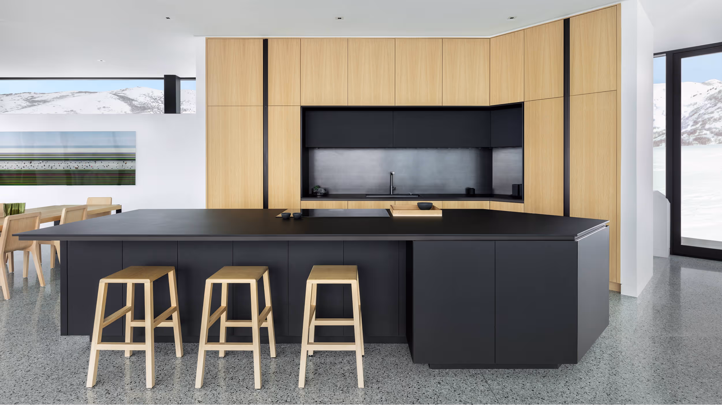 Modern kitchen with black island, wood stools, and wood cabinets under large windows showing snowy mountains.