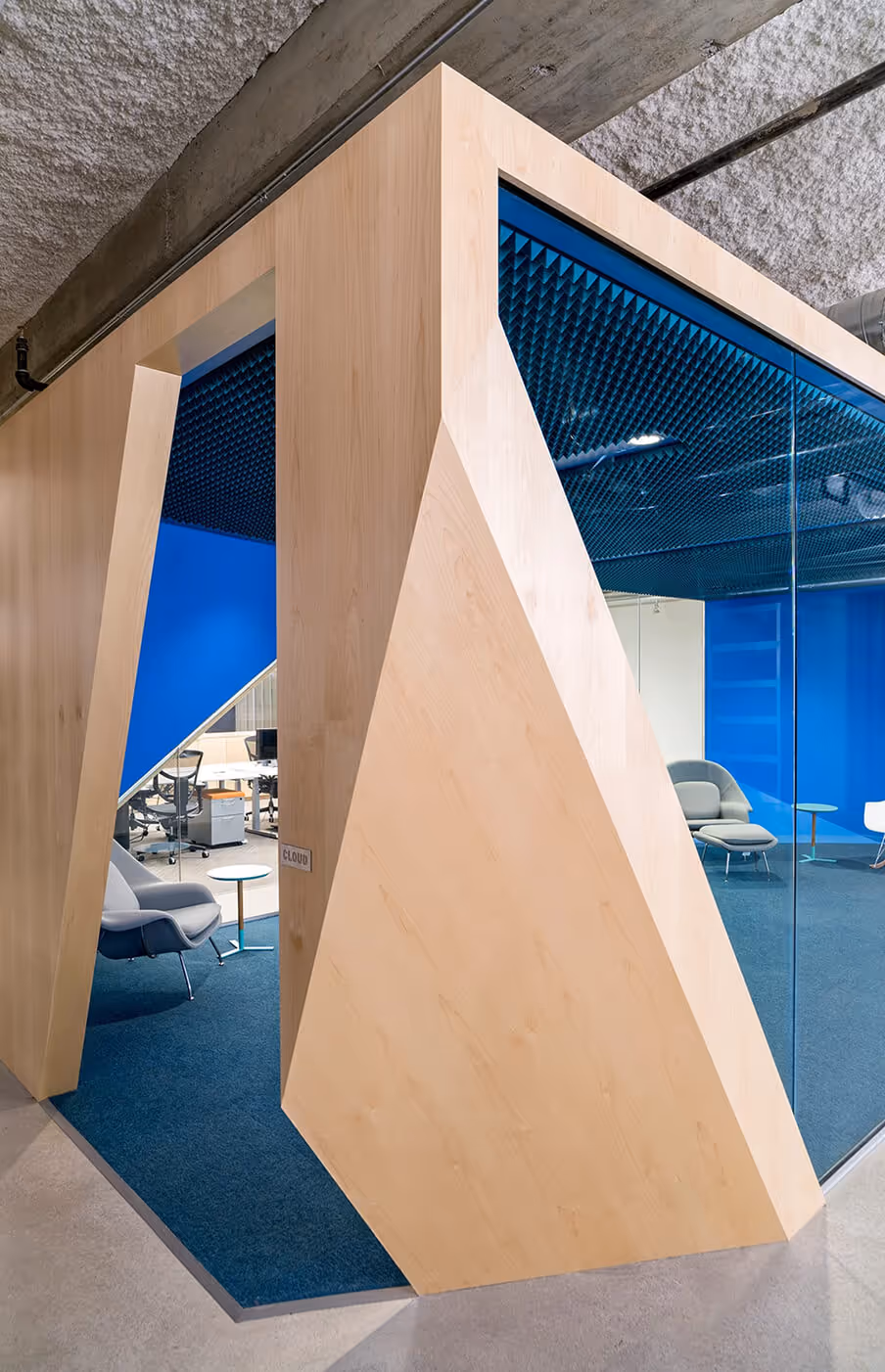 Modern office space with geometric wooden room divider, blue carpet, and gray lounge chairs.
