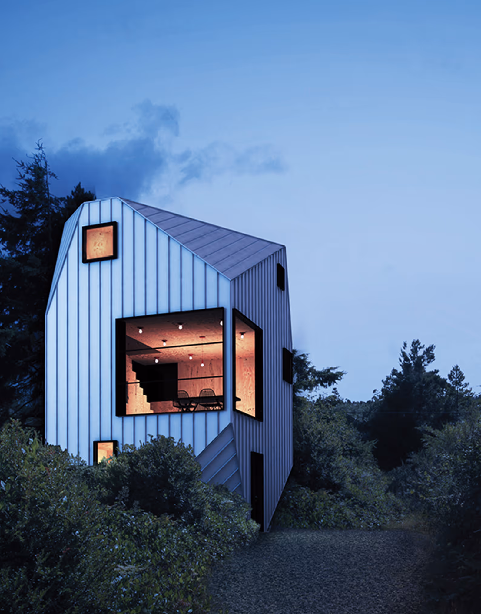 Modern narrow house with large corner windows glowing warmly in wooded area at dusk.