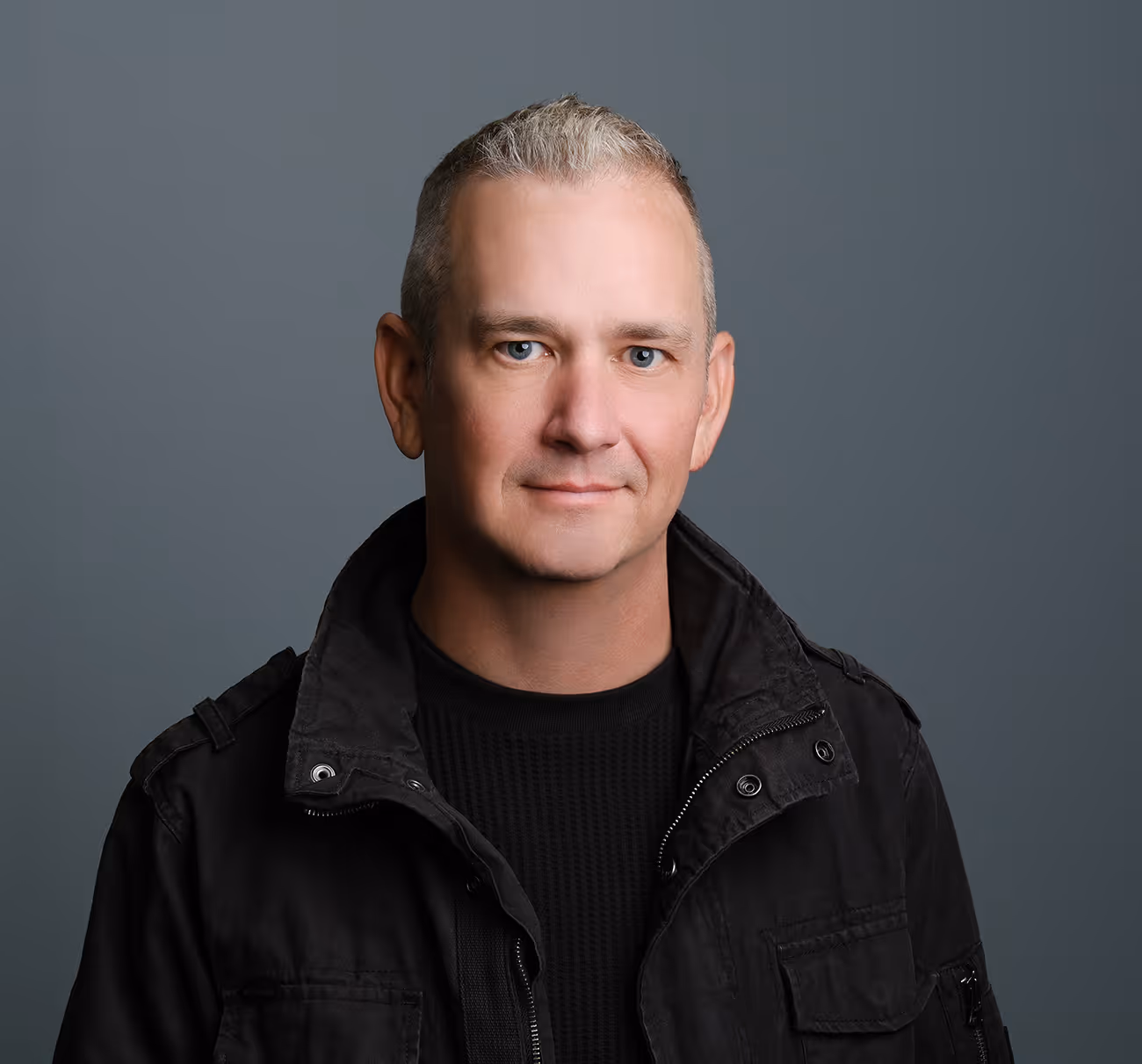 Portrait of a man with short gray hair wearing a black jacket and black sweater against a gray background.