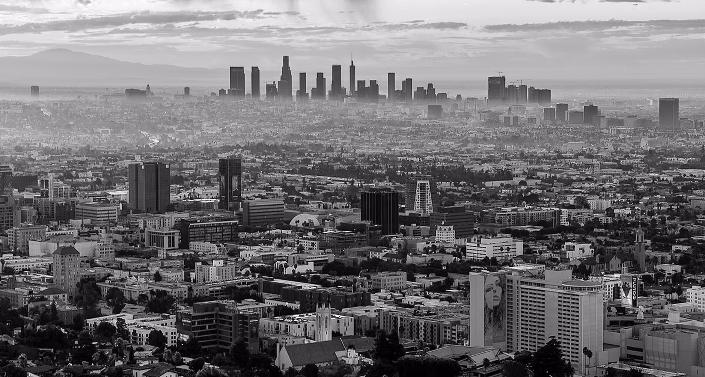 Black and white aerial view of a sprawling cityscape with skyscrapers in the distance under a cloudy sky.