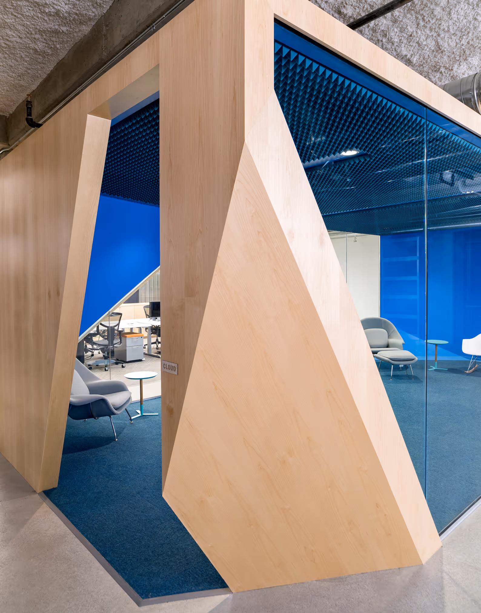 Modern office space with geometric wooden walls, blue accents, and glass partitions containing chairs and small tables.