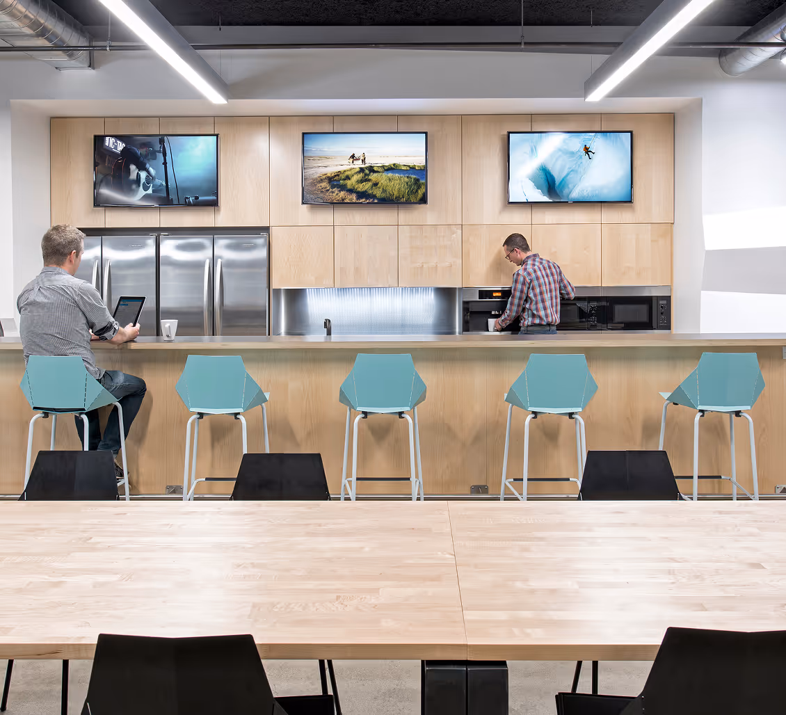 Modern office kitchen with light wood finishes, a long counter with blue stools, and two men using devices.