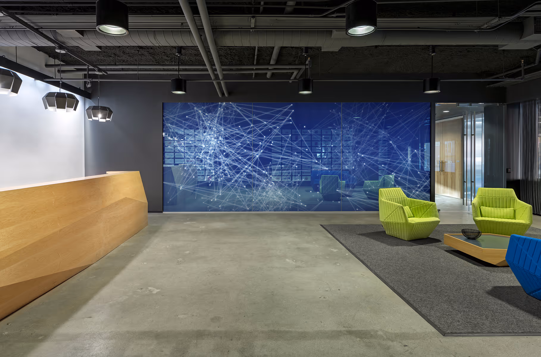 Modern office lobby with wood reception desk, digital network wall art, and green and blue lounge chairs.