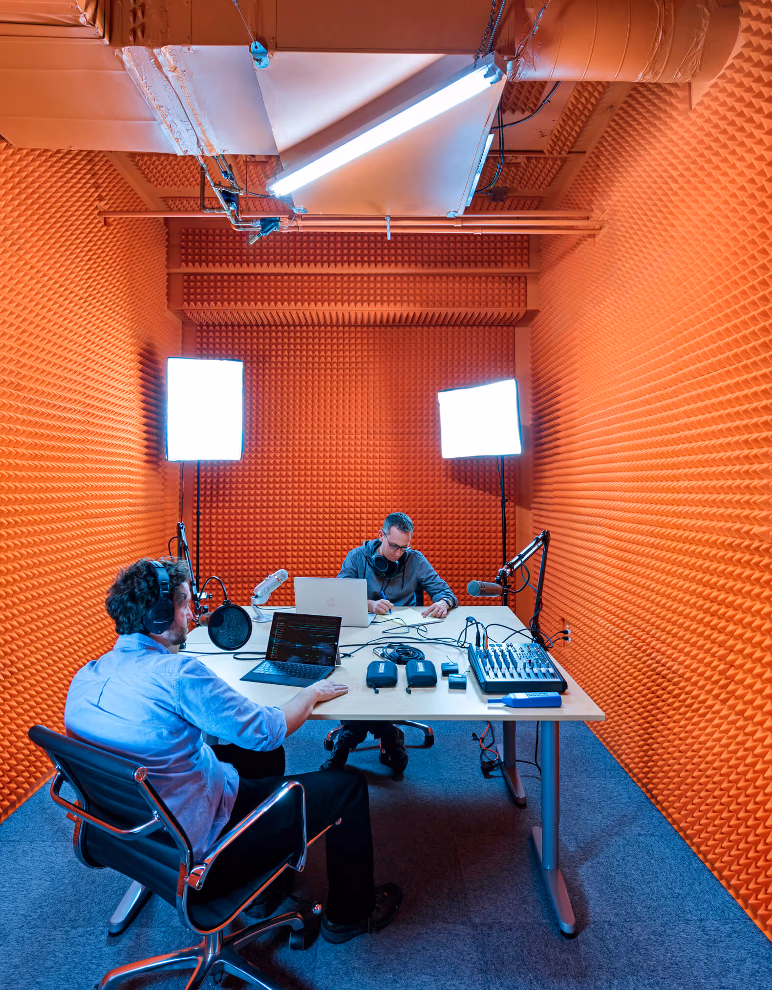 Two men in an orange soundproof room with microphones, laptops, and audio equipment recording a podcast.