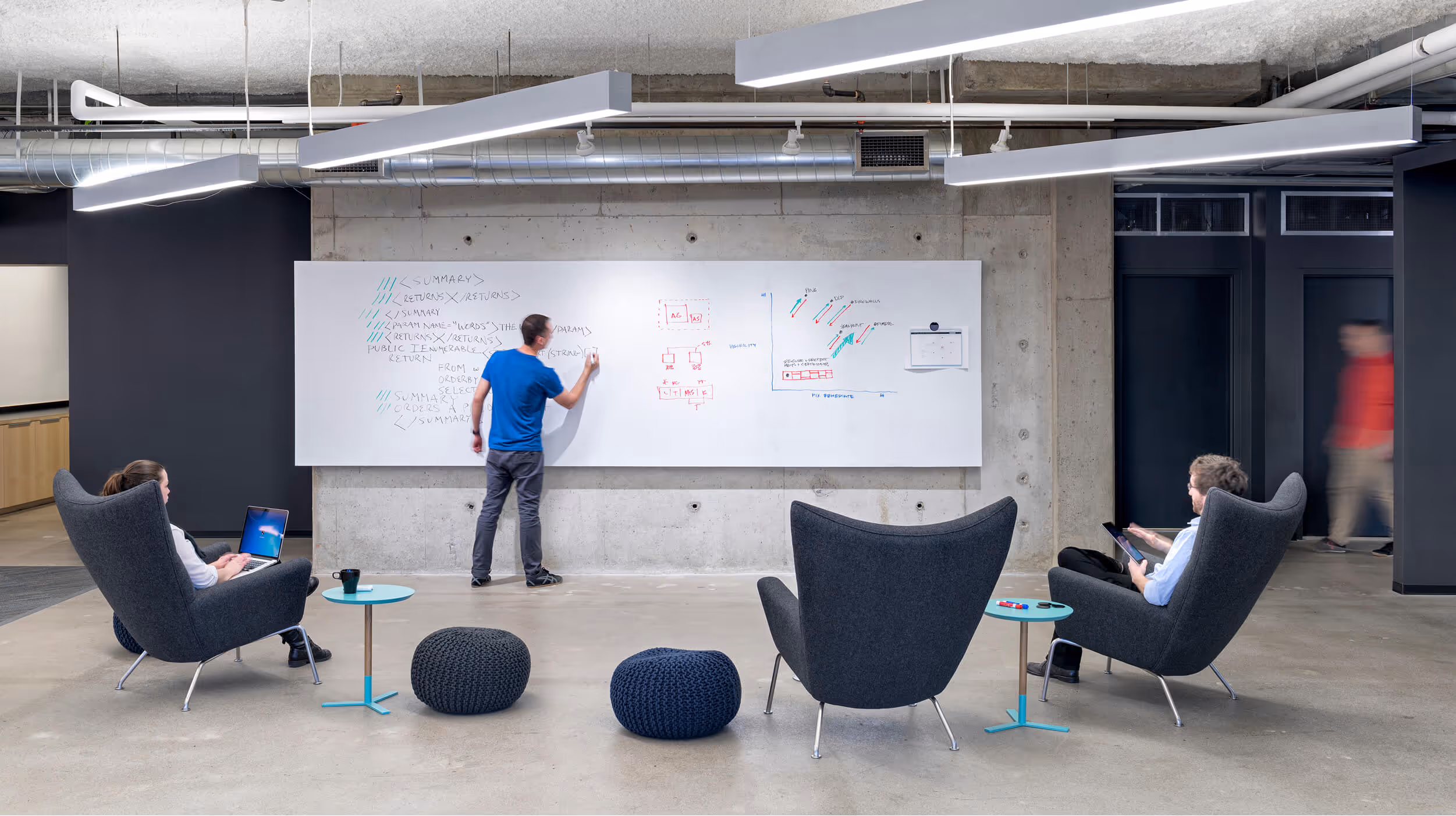 Man writing code on a whiteboard while two others use devices seated in modern office chairs.
