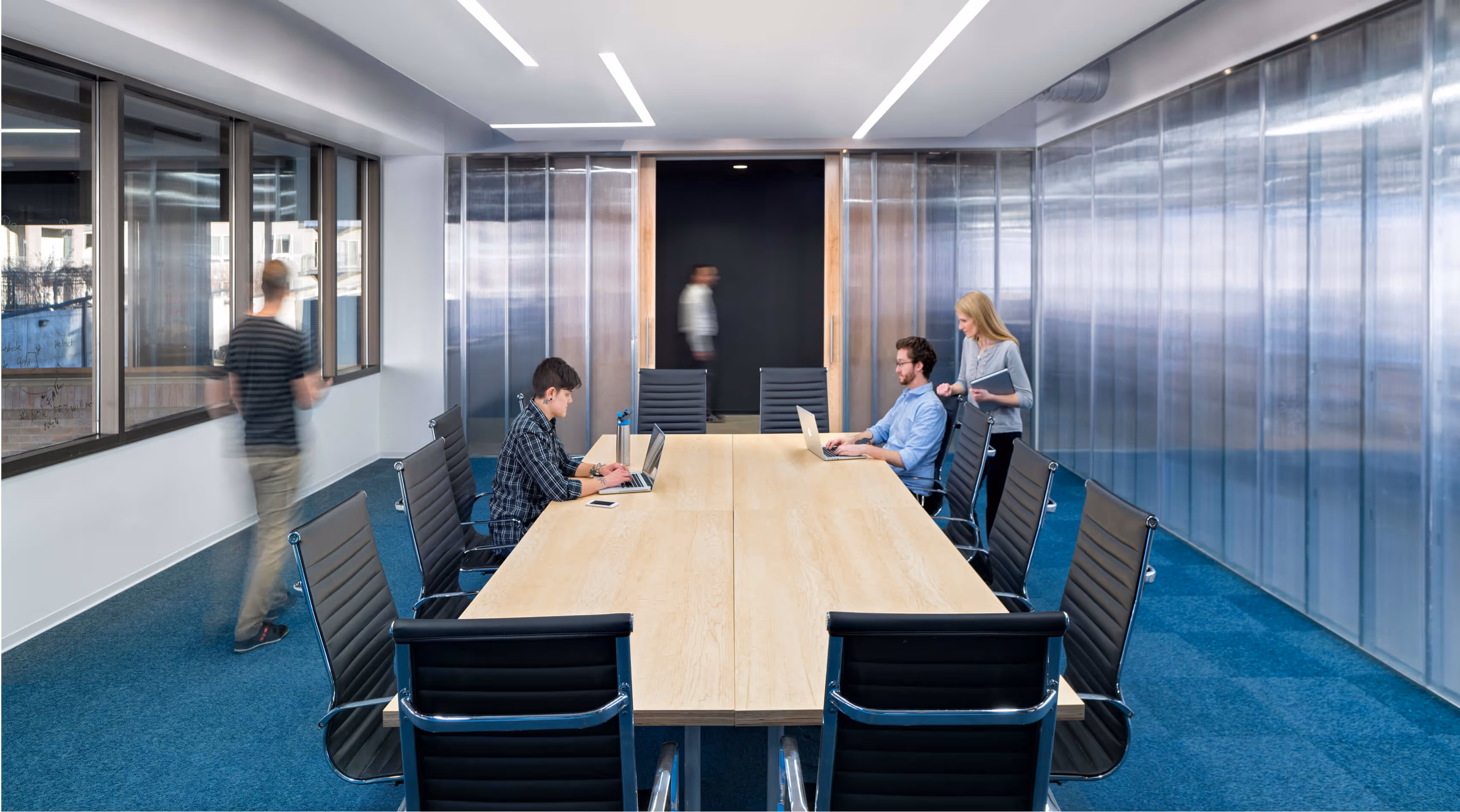 Modern conference room with a long table, black chairs, and people working on laptops.