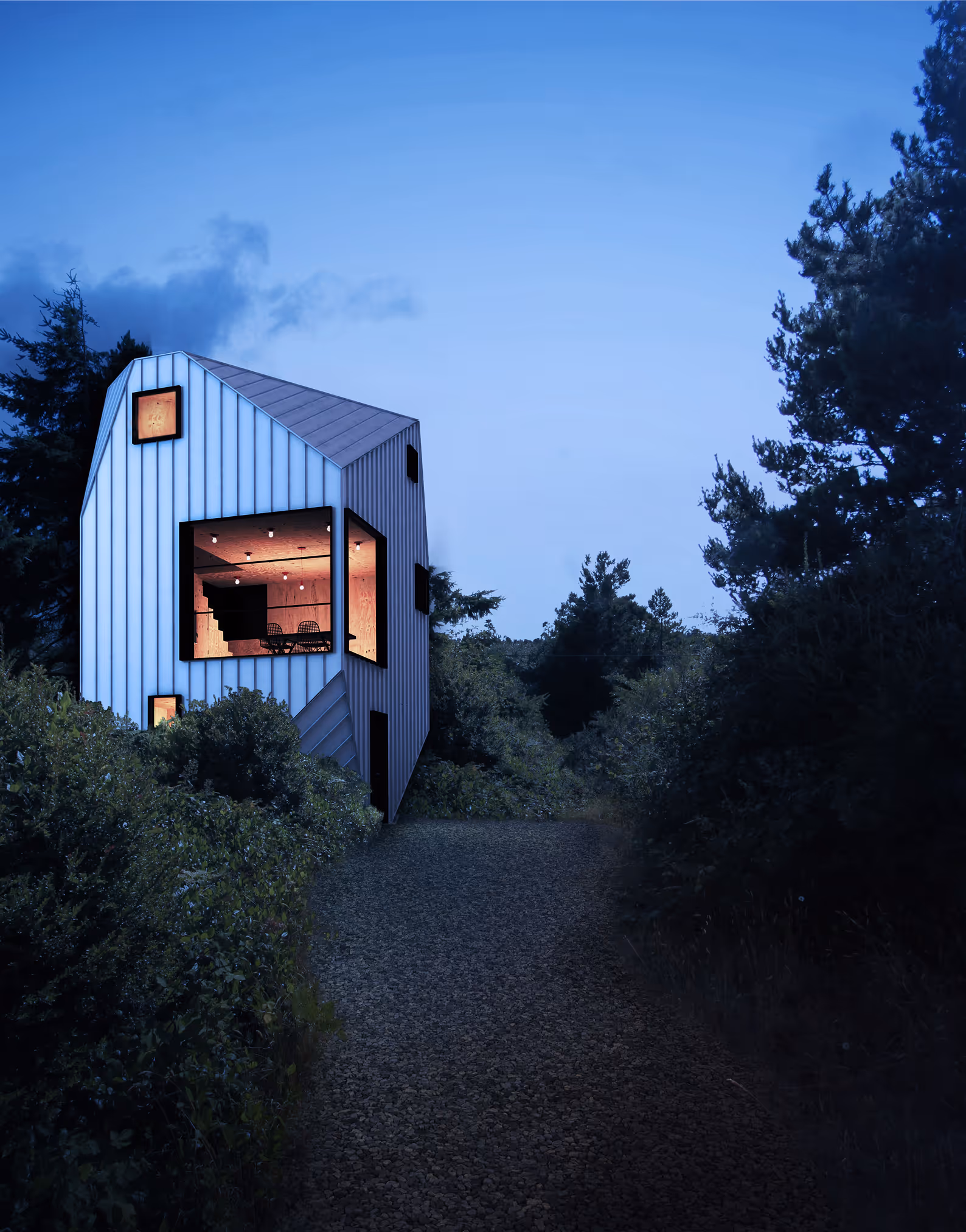 Modern house with illuminated large corner window surrounded by trees at dusk on a gravel path.
