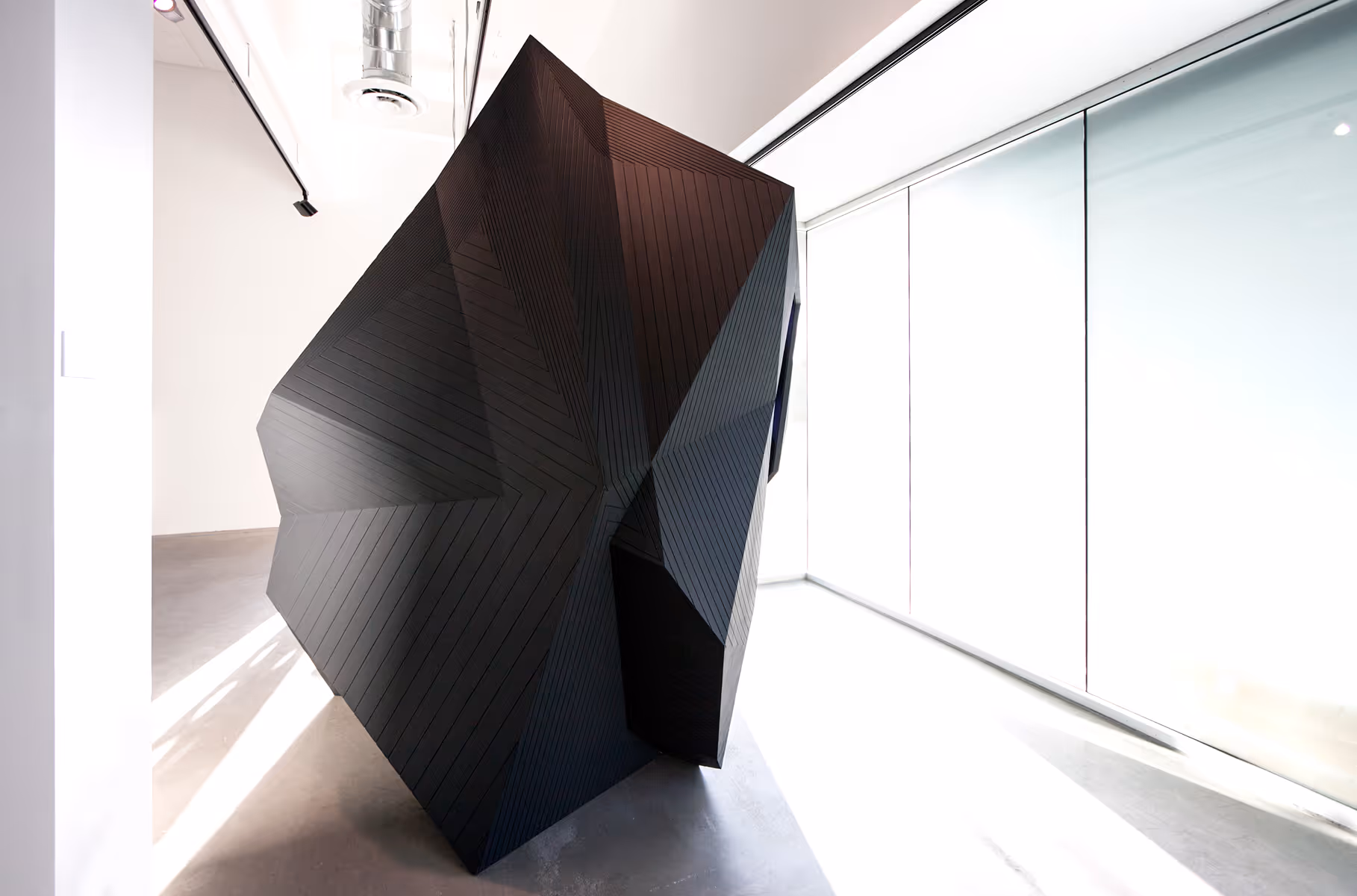 Large black geometric sculpture with angular facets inside a bright modern gallery space.
