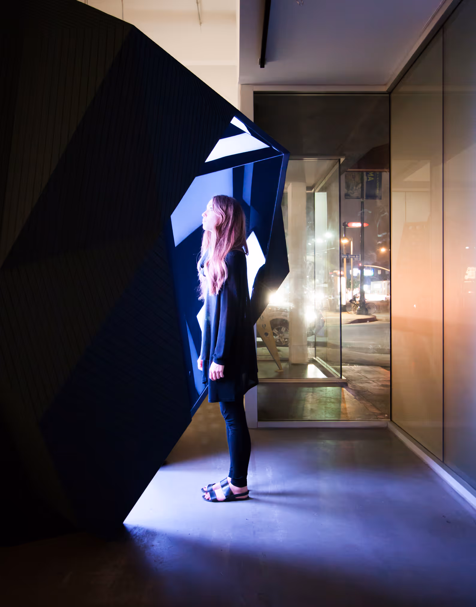 Woman with long hair stands inside a modern art installation glowing with blue light at night.