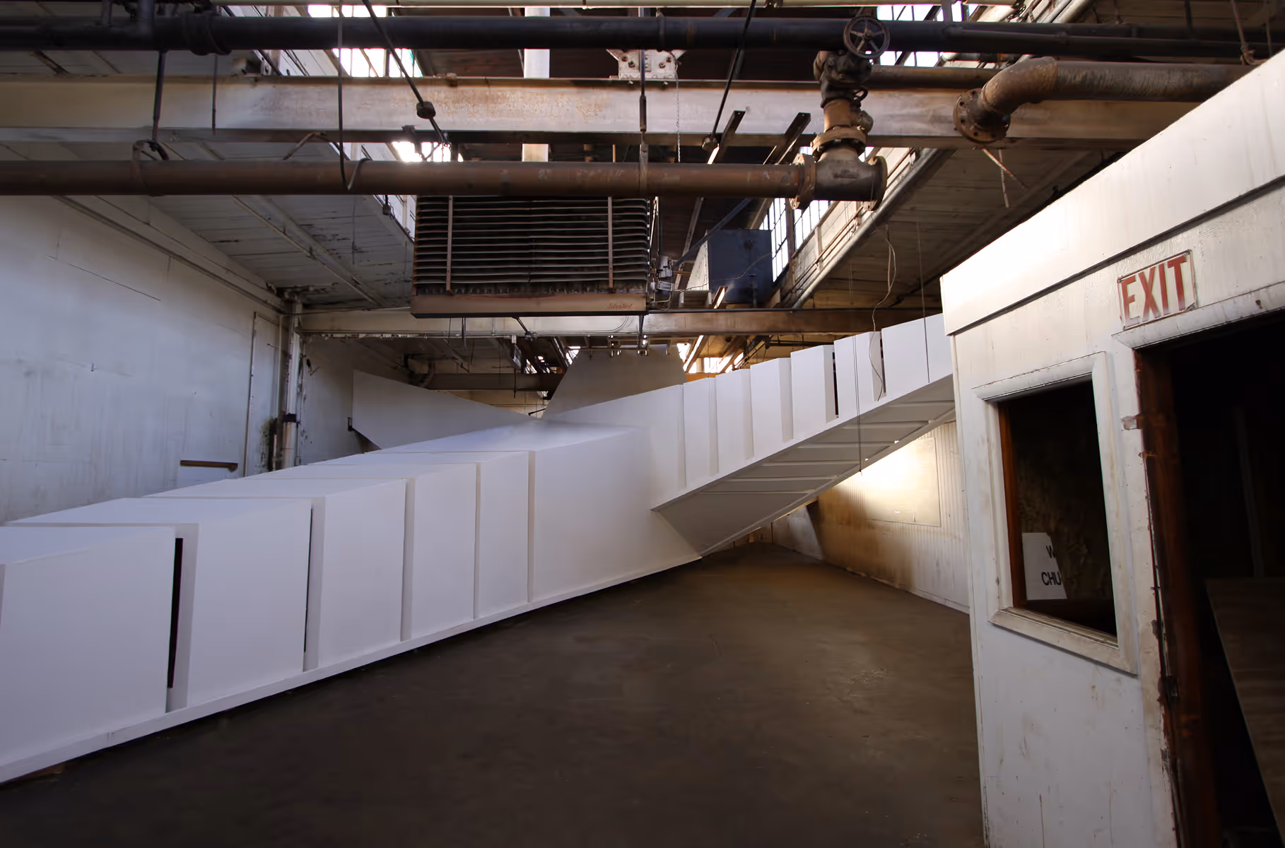 Large white folded rectangular structure inside industrial space with exposed pipes and an exit door.