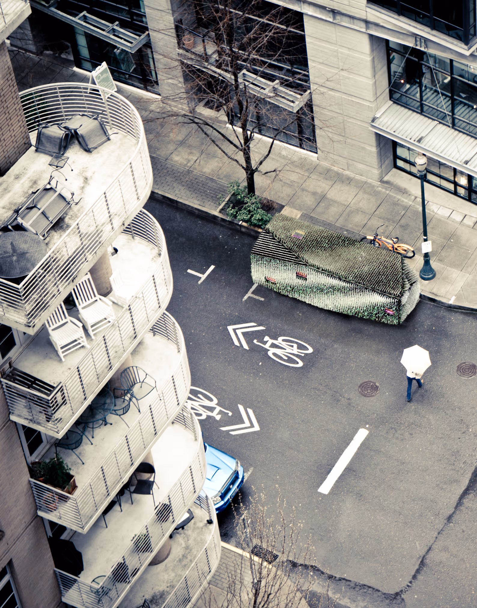 Aerial view of curved balconies on building, bike lane markings, a person with umbrella, and parked blue car.