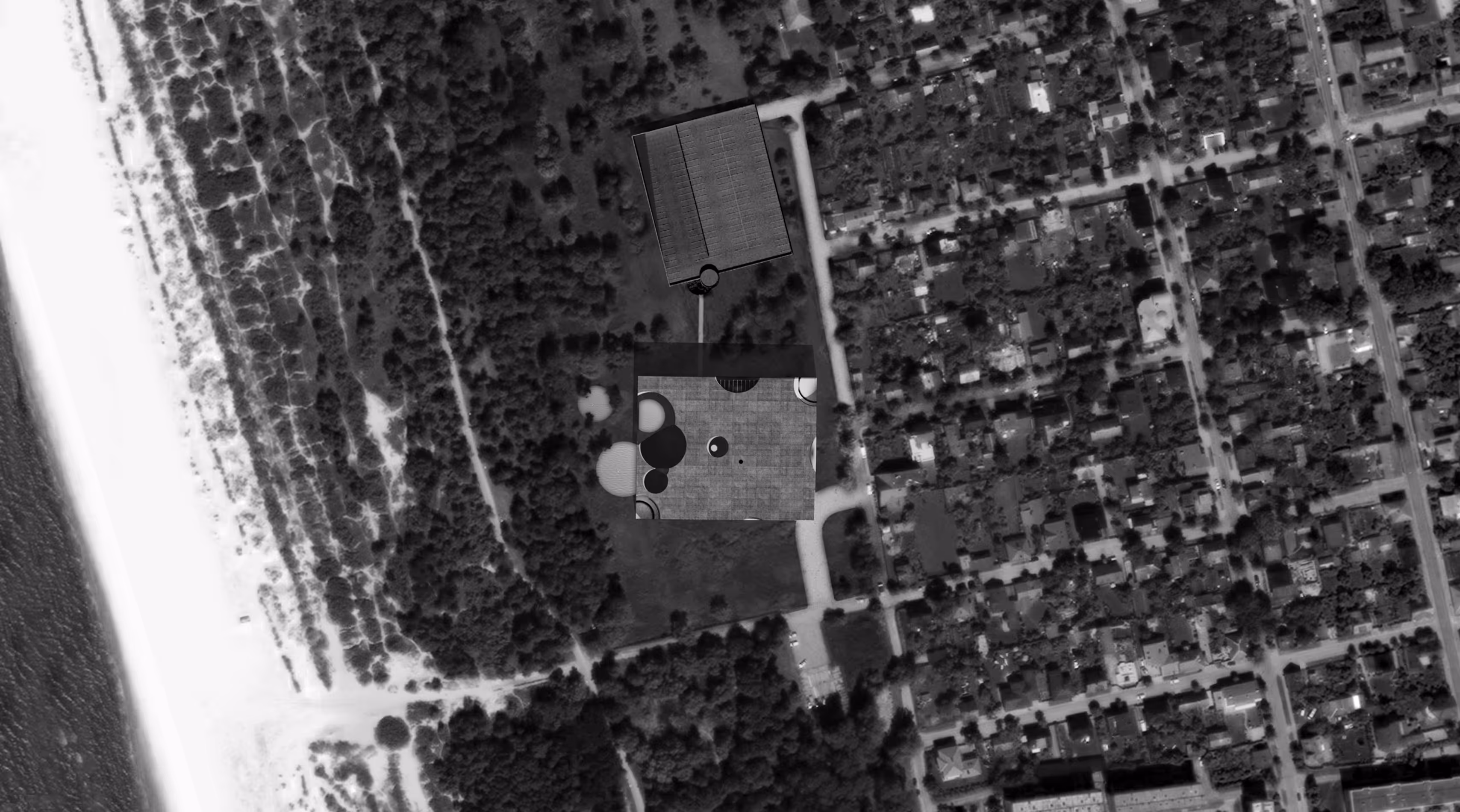 Black and white aerial view of coastal area with beach, forest, a building, and residential neighborhood.